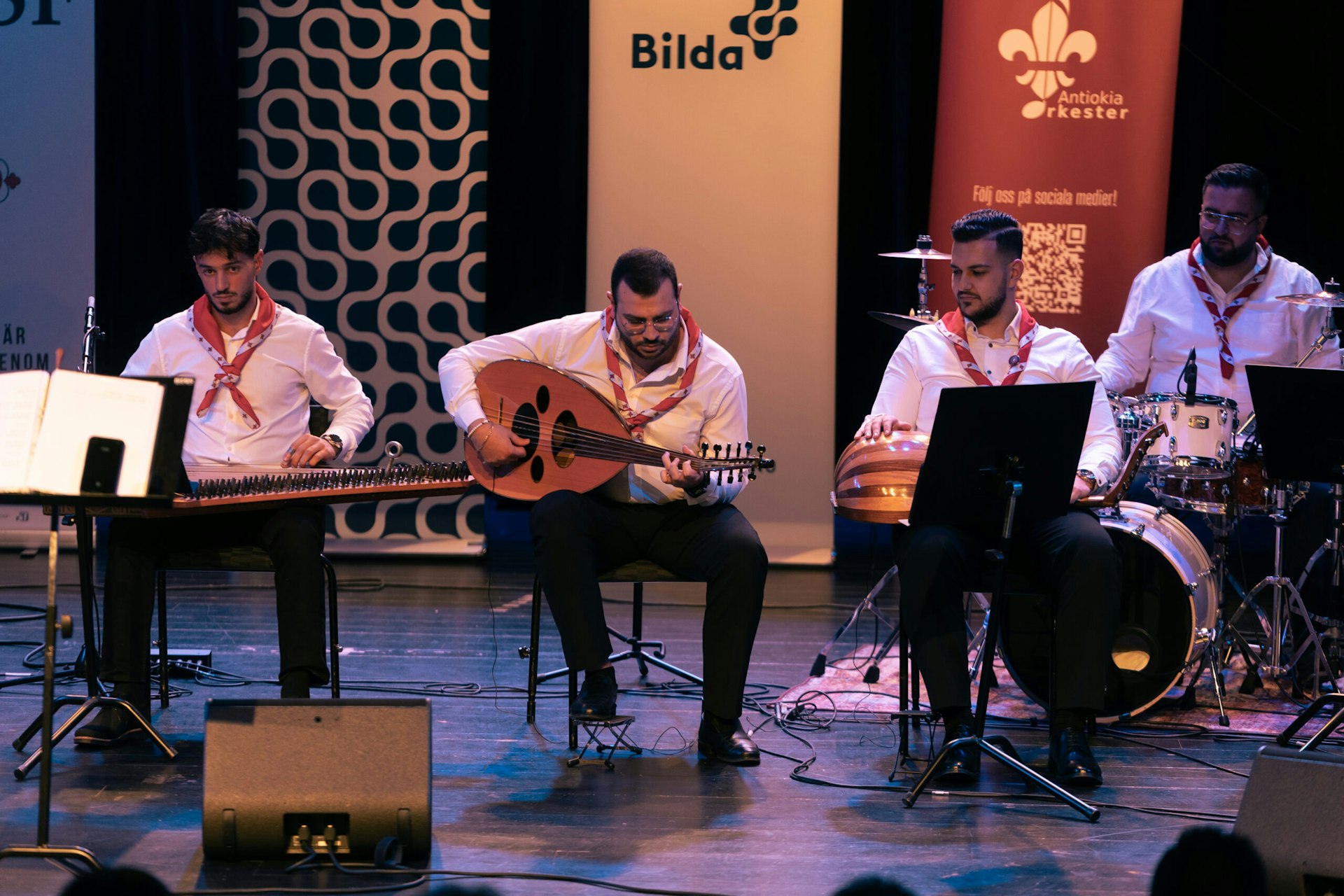 Fyra musiker i vita skjortor uppträder på scenen med traditionella instrument från Mellanöstern och ett trumset.