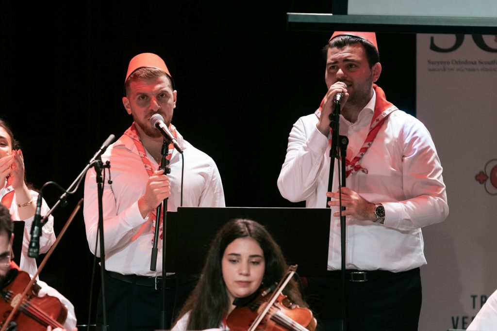 Två män i vita skjortor och röda hattar sjunger i mikrofoner och framför dem spelar en violinist.