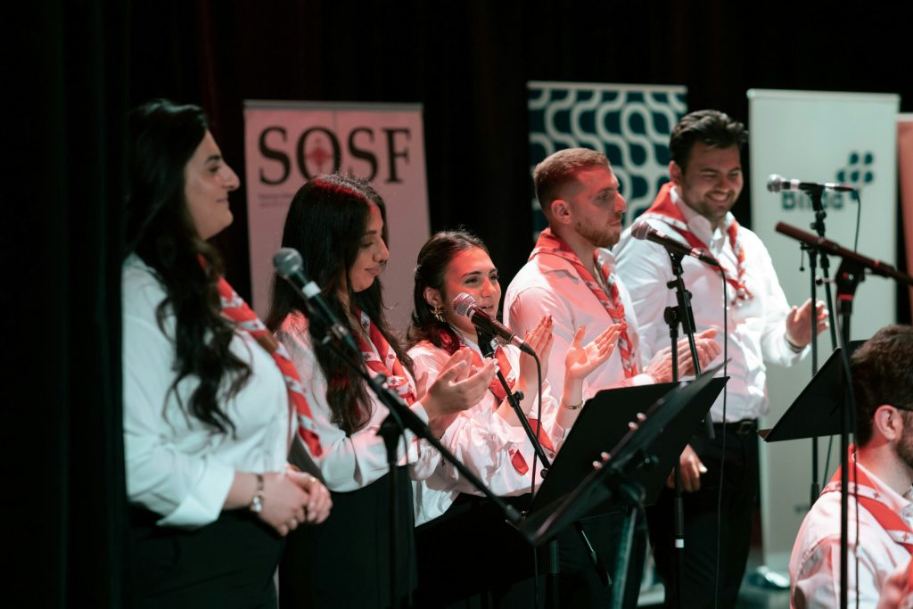 Fem personer i vita skjortor och röda halsdukar sjunger och gestikulerar på scenen med mikrofoner och notställ.