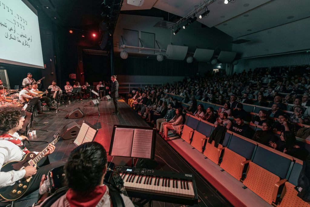Ett band uppträder på scenen inför en stor sittande publik i ett inomhusauditorium.