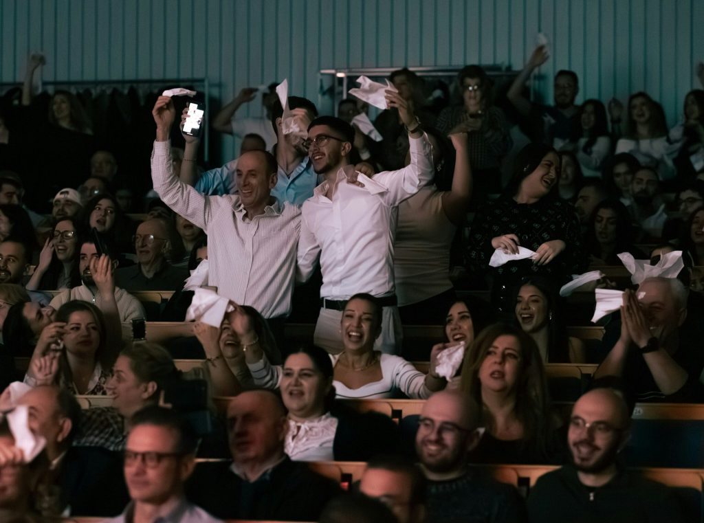 Publiken i en teater jublar och viftar med näsdukar, många människor ler och står upp i spänning.
