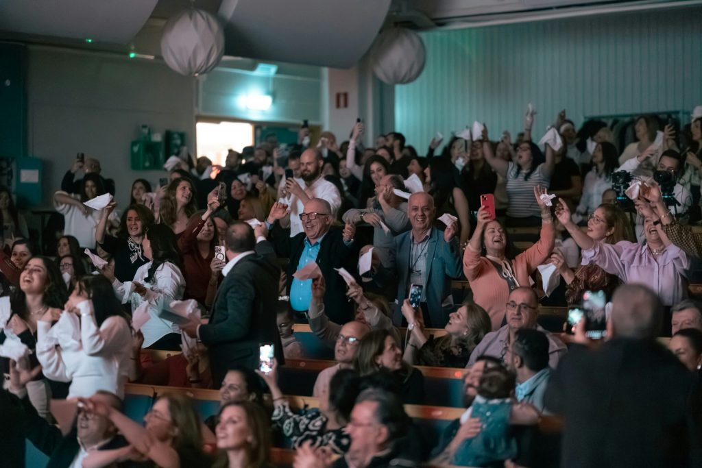En stor publik i ett auditorium viftar med vita papper och ler, många tar bilder med sina telefoner.