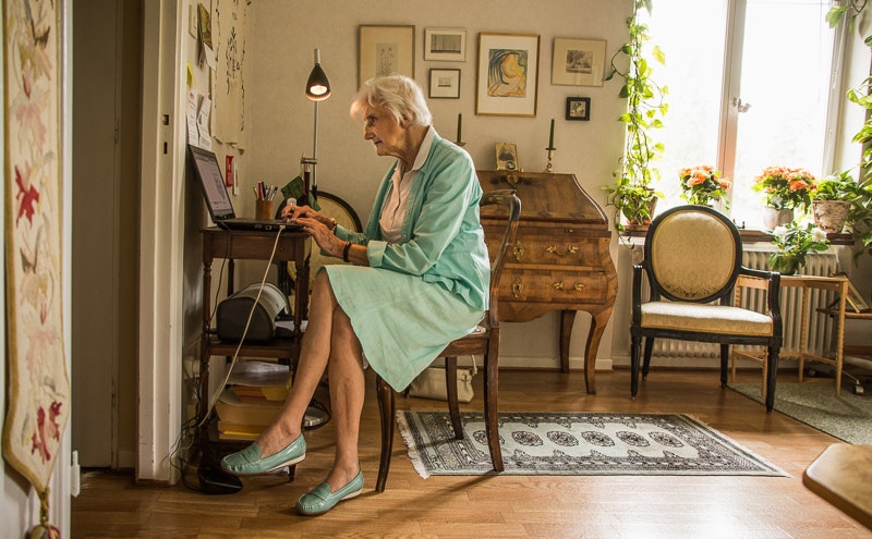 Äldre kvinna i blågrön klänning använder en bärbar dator vid ett skrivbord i ett mysigt, solbelyst vardagsrum med växter och konstverk.