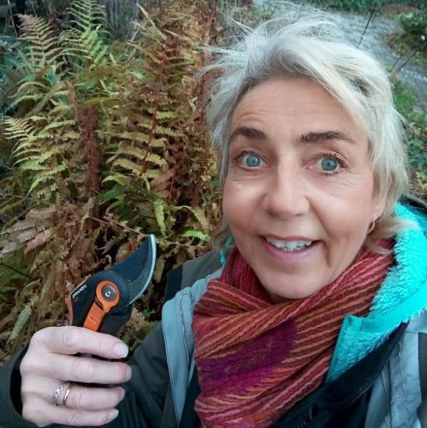 Leende kvinna i röd halsduk håller i en sekatör bredvid bruna ormbunkar i en trädgård.