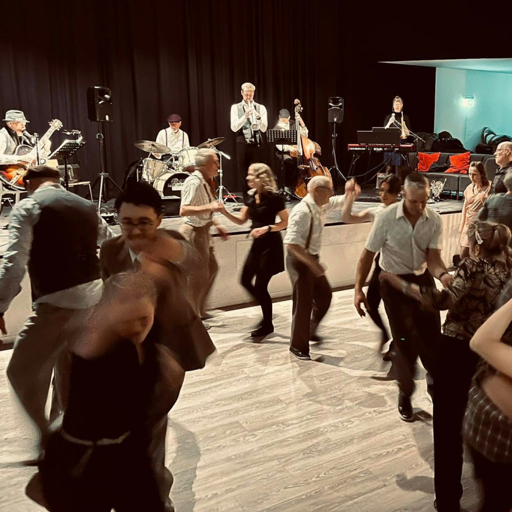 Människor dansar swingdans i par på ett dansgolv med ett liveband som spelar på scenen i bakgrunden.