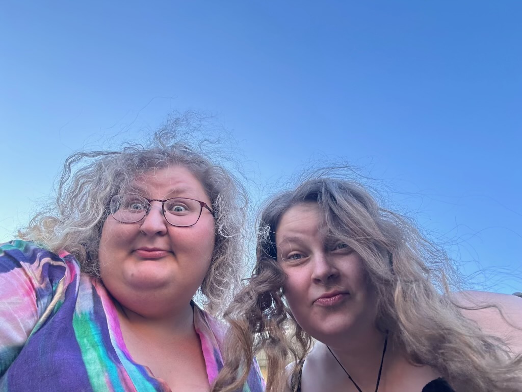 Två kvinnor med lockigt hår gör fåniga miner mot kameran, mot en klarblå himmel.