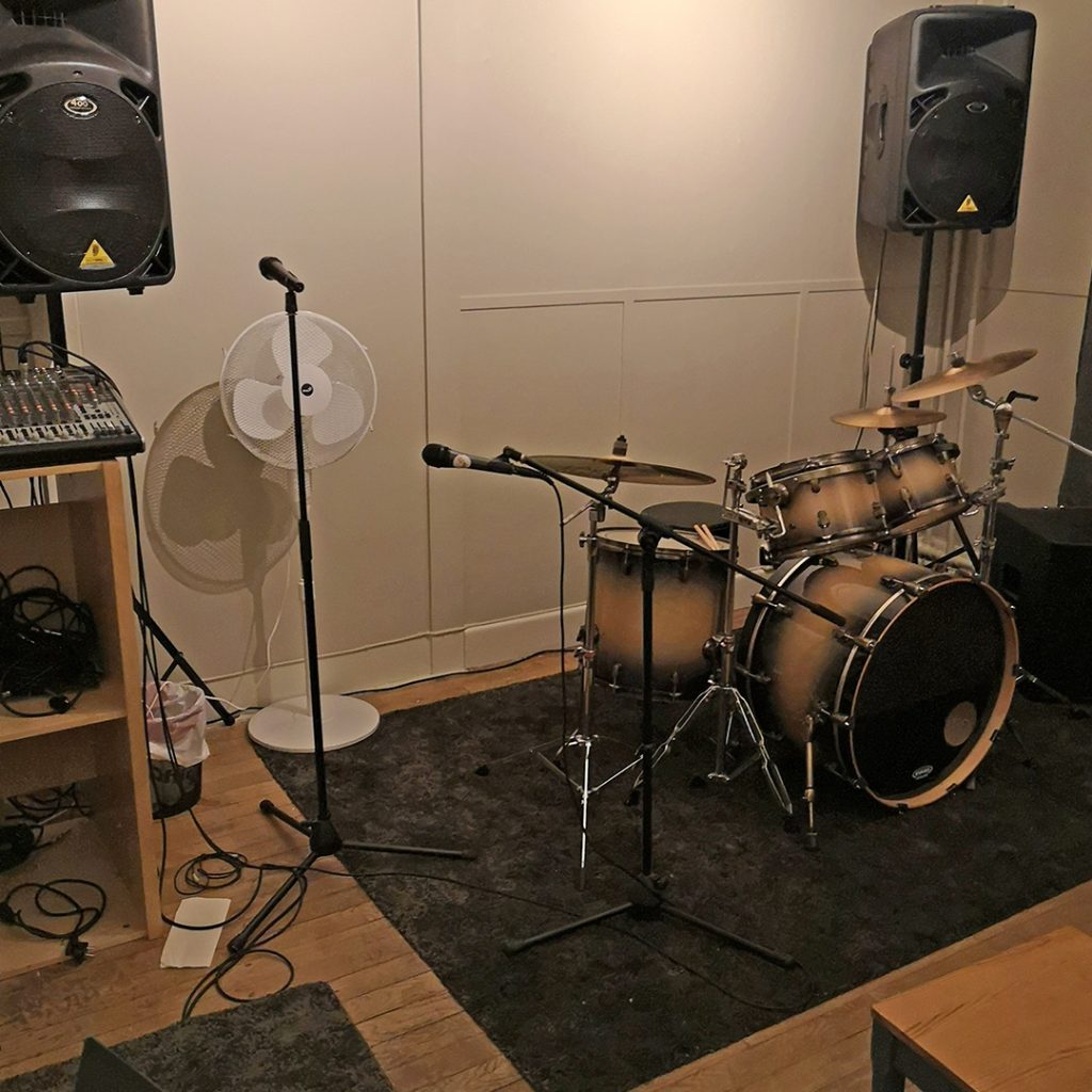 Ett trumset, mikrofoner, högtalare och en fläkt i ett musikrepetitionsrum med trägolv.