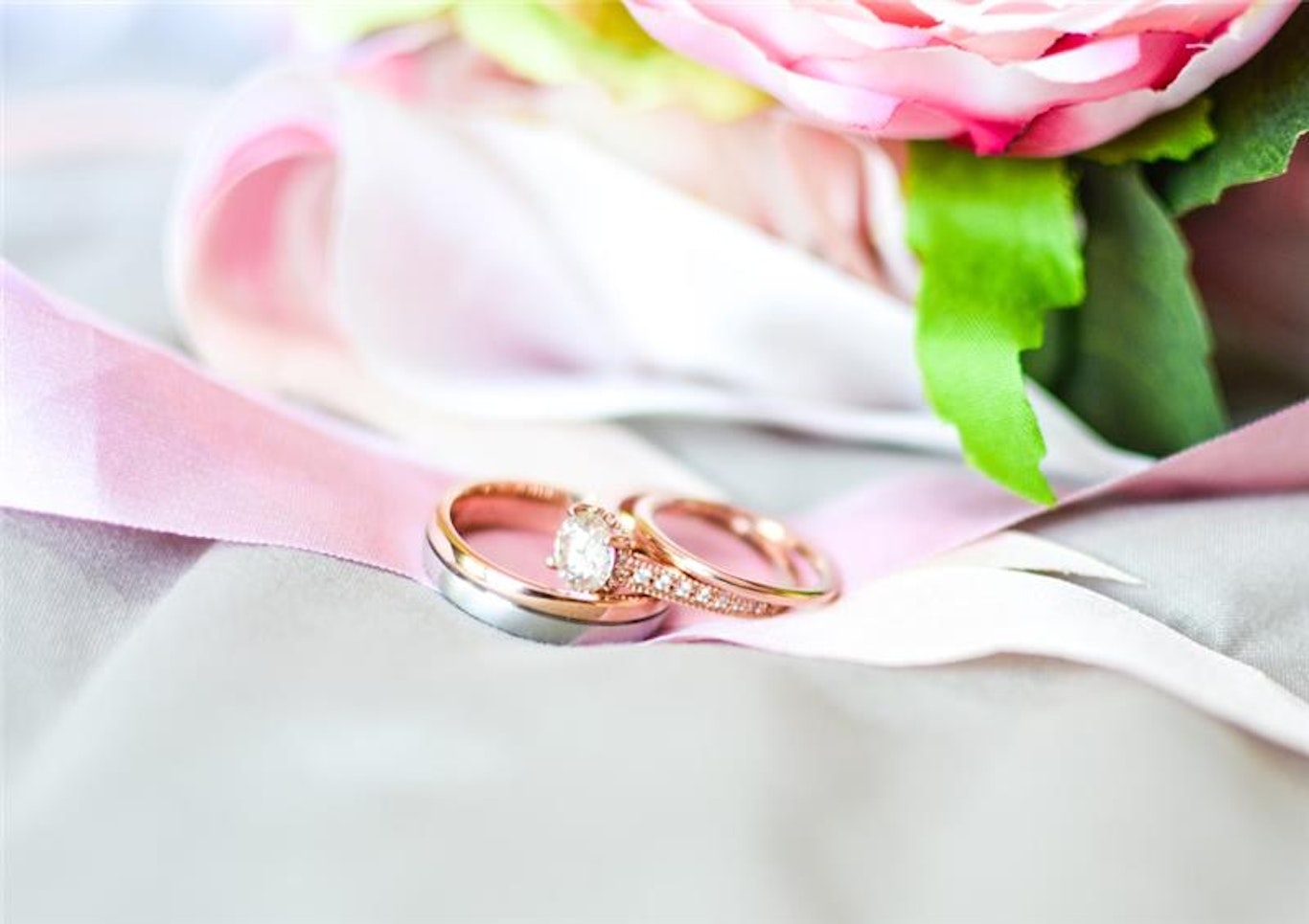 Två vigselringar på rosa och vitt band med en rosa och vit blomma i bakgrunden.