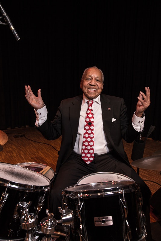 En äldre man i kostym och rödmönstrad slips ler när han sitter vid ett trumset på scenen.