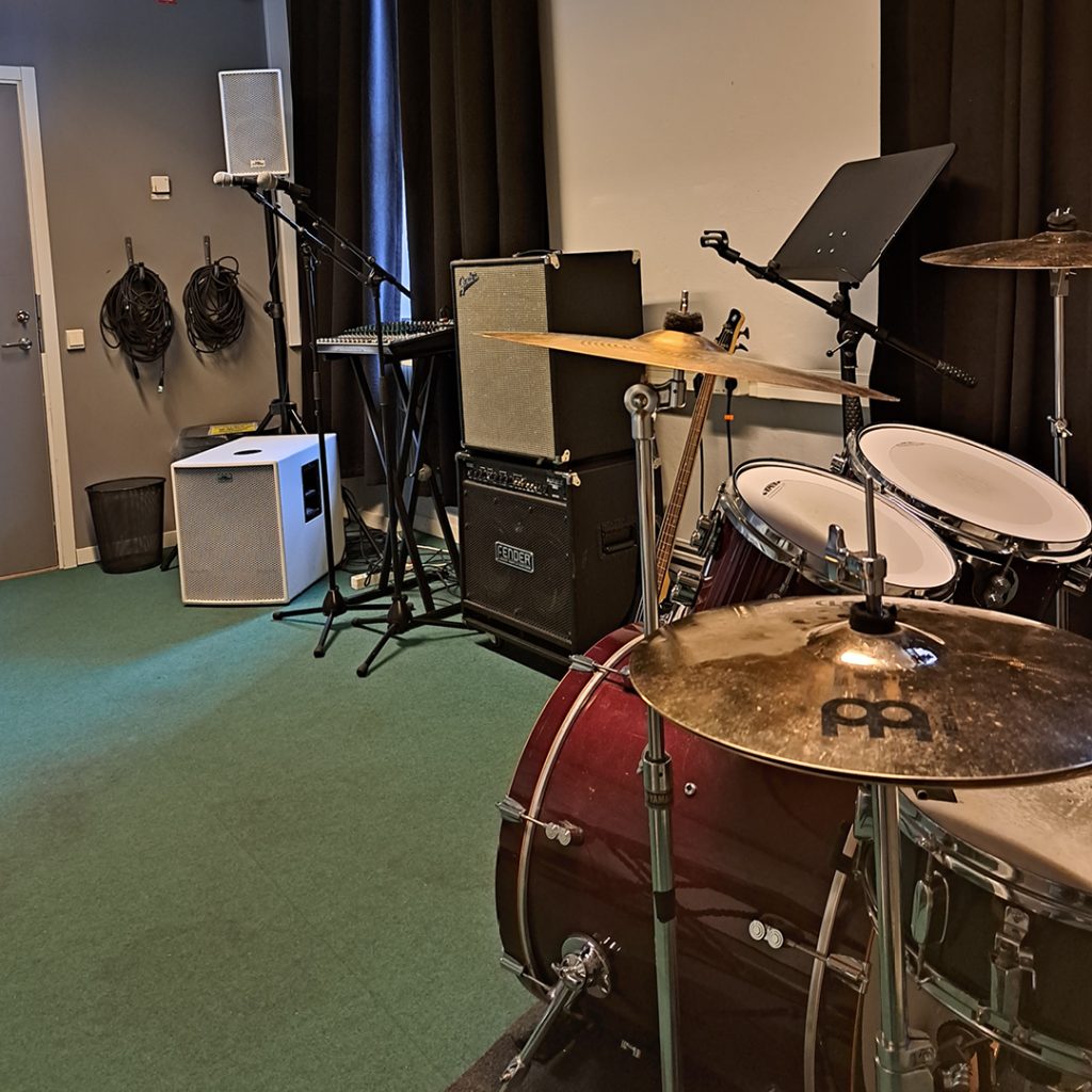Ett musikrepetitionsrum med trumset, förstärkare, keyboard och notställ på en grön matta.