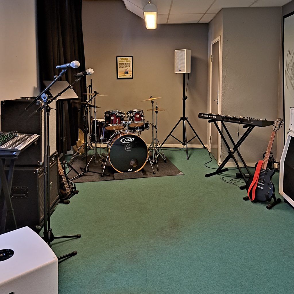 En replokal för musik med trummor, keyboard, gitarr, högtalare och mikrofoner på stativ.