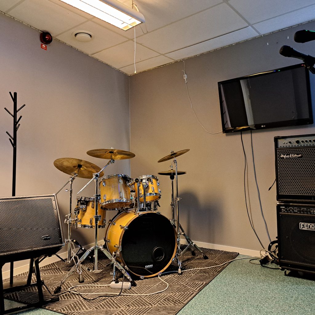 Ett gult trumset, högtalare och ett mikrofonstativ i ett litet musikövningsrum med en TV på väggen.