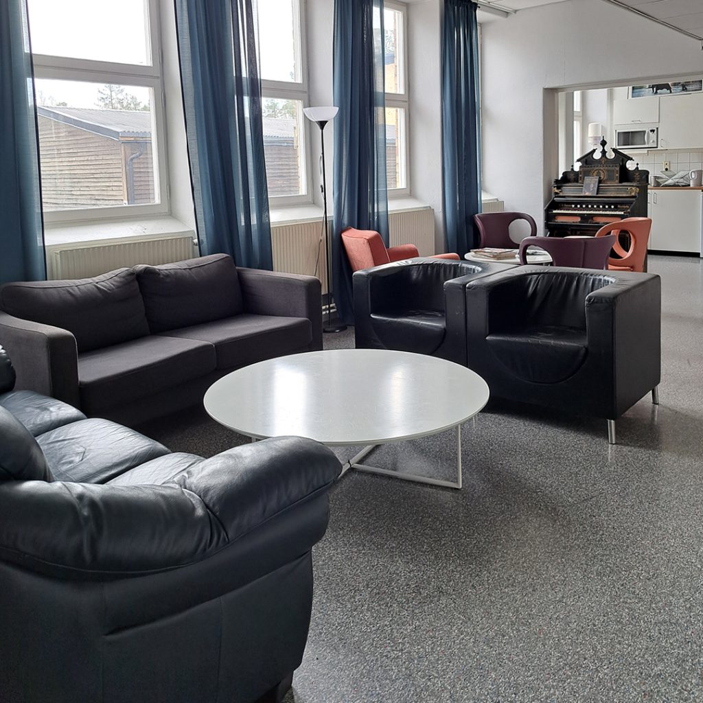 Modernt vardagsrum med svarta soffor, runt vitt bord, köksdel och stora fönster med blå gardiner.