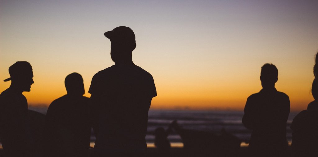 Silhuetter av människor som står på en strand i solnedgången, med en orange och lila himmel i bakgrunden.