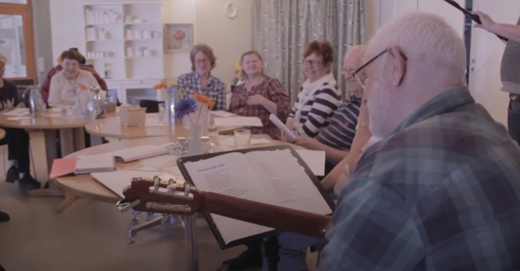 En grupp äldre vuxna sitter runt bord och sjunger tillsammans medan en person spelar gitarr - de njuter av musikglädje och gemenskap genom Vi sjunger hjärna.