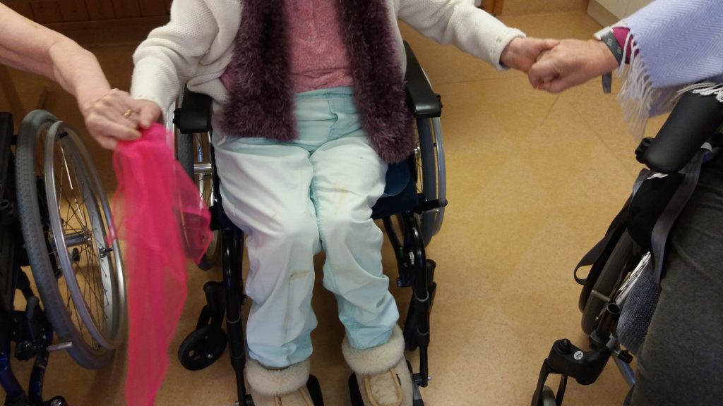 Tre personer håller varandra i handen i en cirkel, två i rullstol, en stående, alla inomhus på ett solbränt golv.