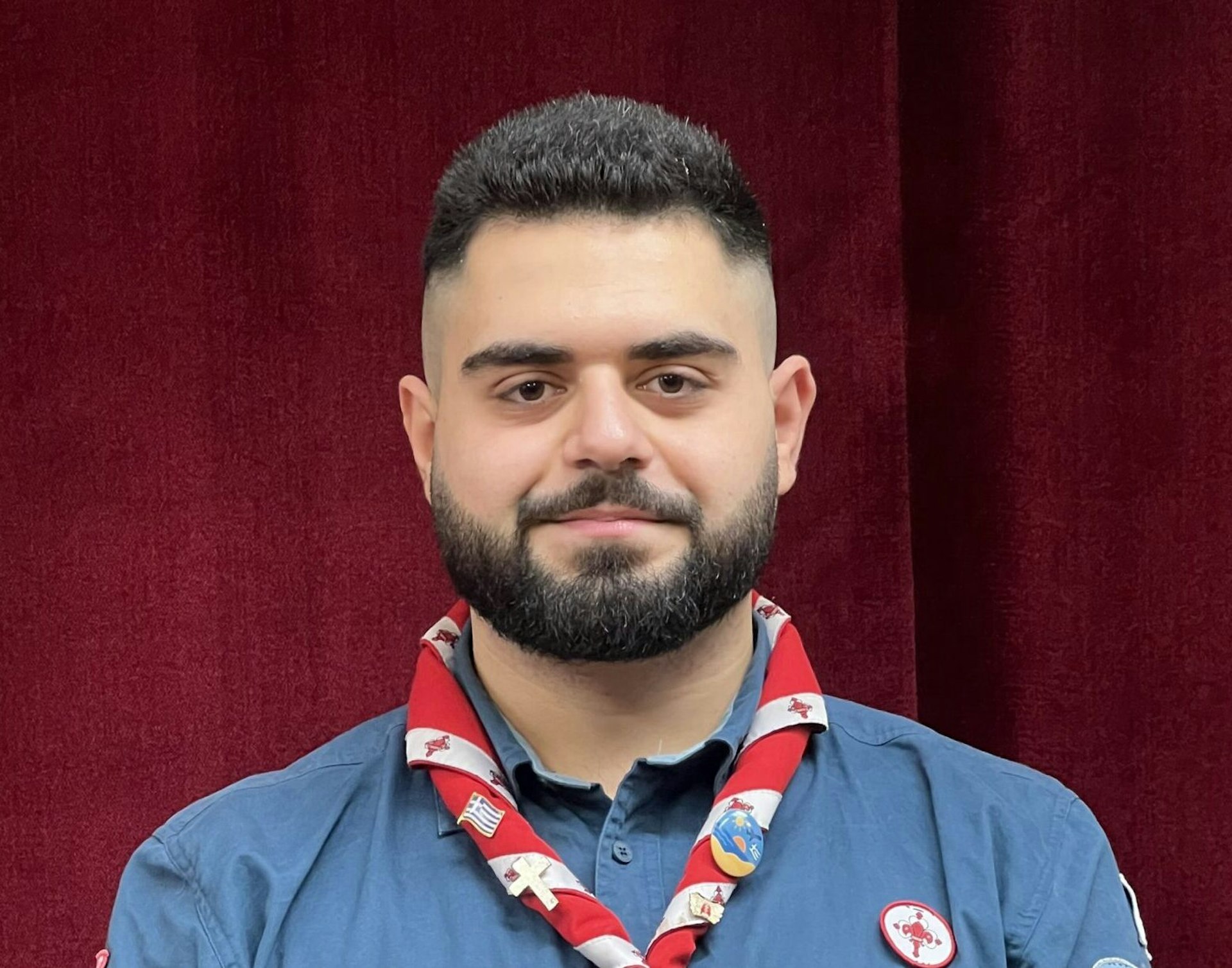 En man med mörkt skägg i scoutuniform står framför en röd ridå och ler lite.