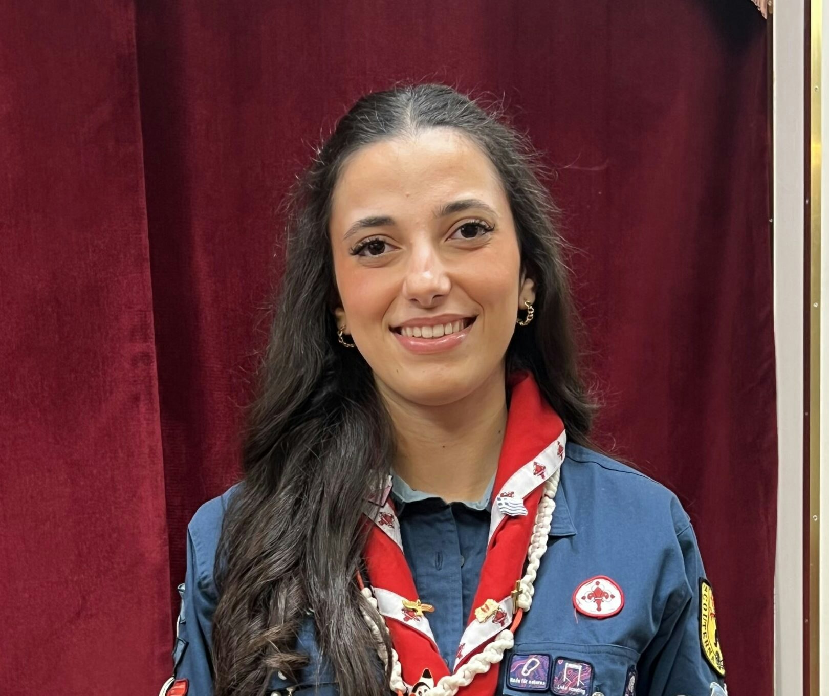 En kvinna i marinblå scoutuniform Scouterna och röd halsduk ler framför en mörkröd ridå.