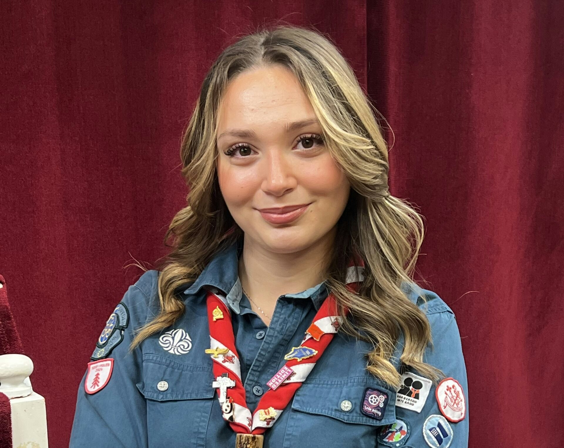 Ung kvinna i blå scoutuniform med märken, står framför en röd gardin och ler mot kameran.