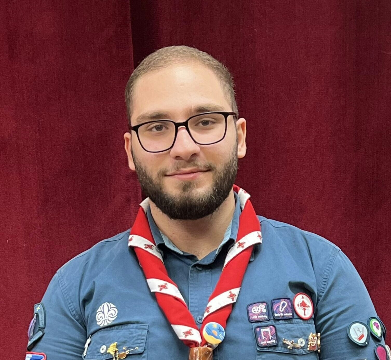 En person med glasögon och scoutuniform från Scouterna står framför en röd ridå och ler lite.