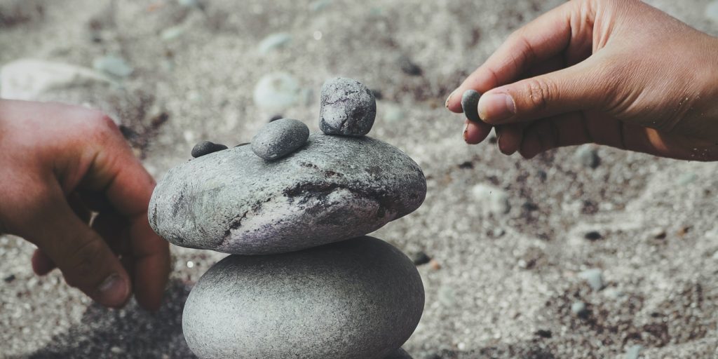 Två händer staplar släta stenar ovanpå varandra på en sandyta.