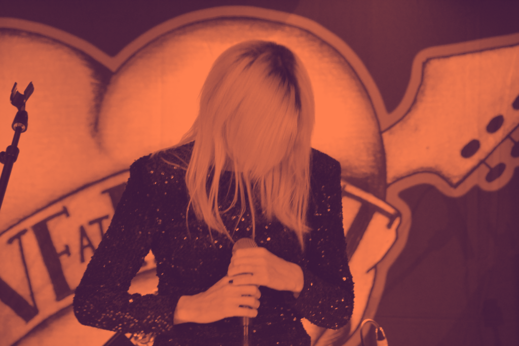 En person i svart paljettdräkt håller en mikrofon med huvudet nedåt och står framför en hjärtformad kuliss.