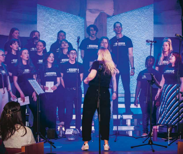 En kör uppträder på scenen under ledning av en kvinna, med blå belysning och mikrofoner omkring sig.