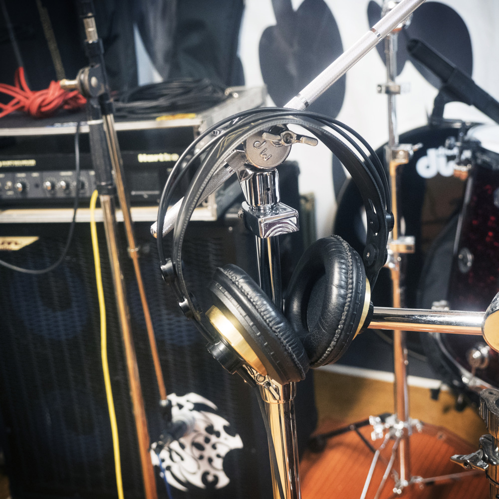 Ett par svarta hörlurar hänger på ett trumstativ i en musikstudio med trummor och förstärkare i närheten.