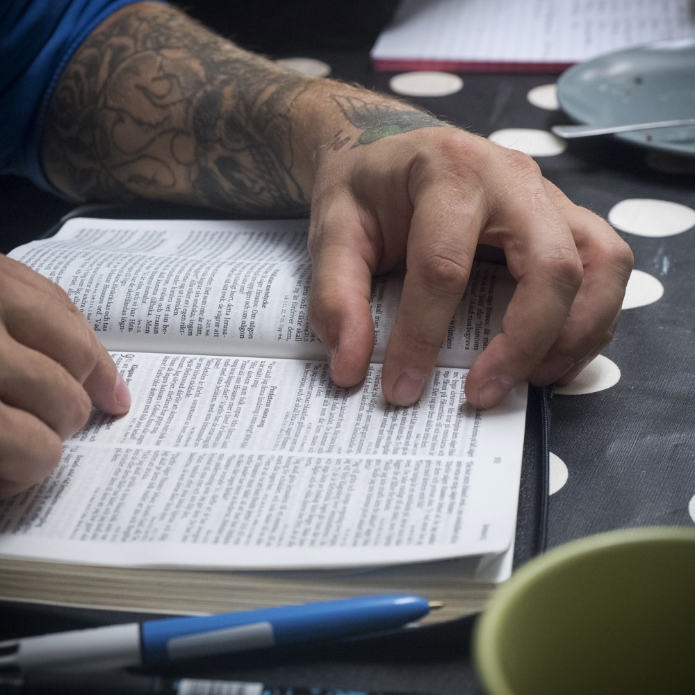 Tatuerade händer håller i och läser en öppen bok på en prickig bordsduk, med en penna och mugg i närheten.
