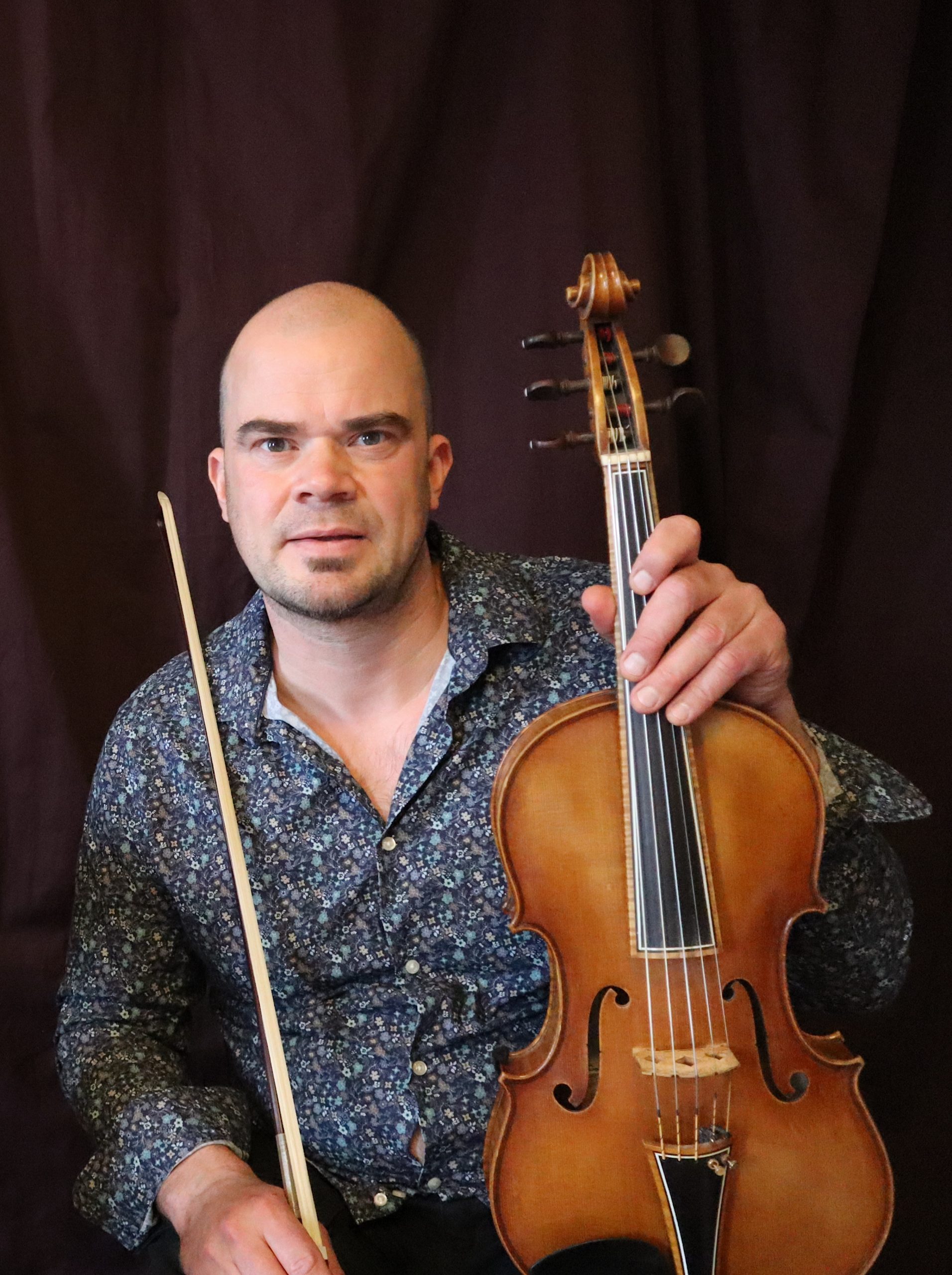 En man i blommig skjorta håller i en viola och en stråke och sitter framför en mörk bakgrund.