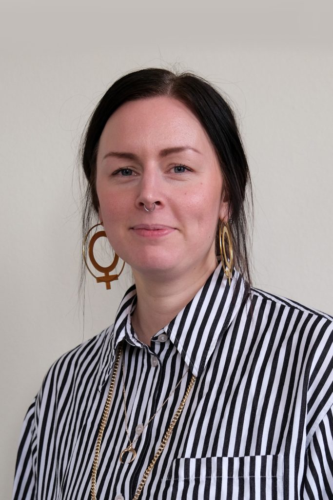 Kvinna med mörkt hår, randig skjorta, stora örhängen och halsband i guld, stående mot ljus bakgrund.