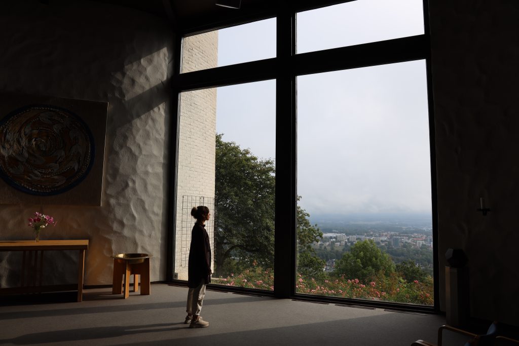 Ett barn står vid ett stort fönster och tittar ut över en stadsvy med träd och blommor i förgrunden.