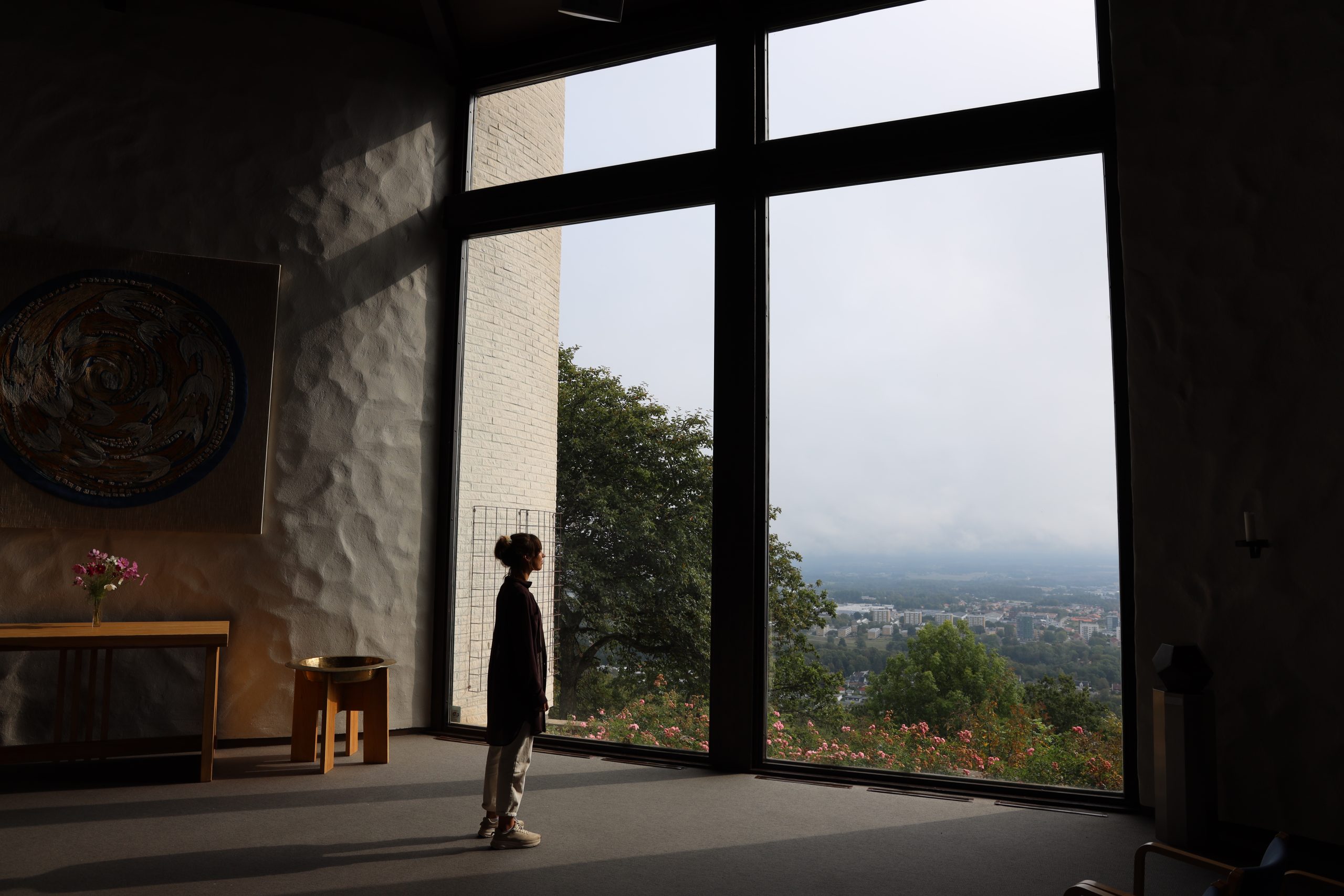 Ett barn står vid ett stort fönster och tittar ut över en stadsvy med träd och blommor i förgrunden.