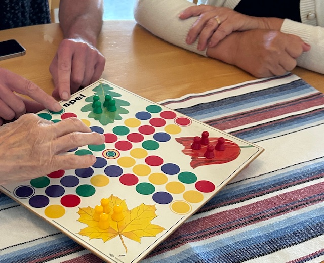 Tre personer spelar ett färgglatt brädspel med bladmönster på en randig duk.