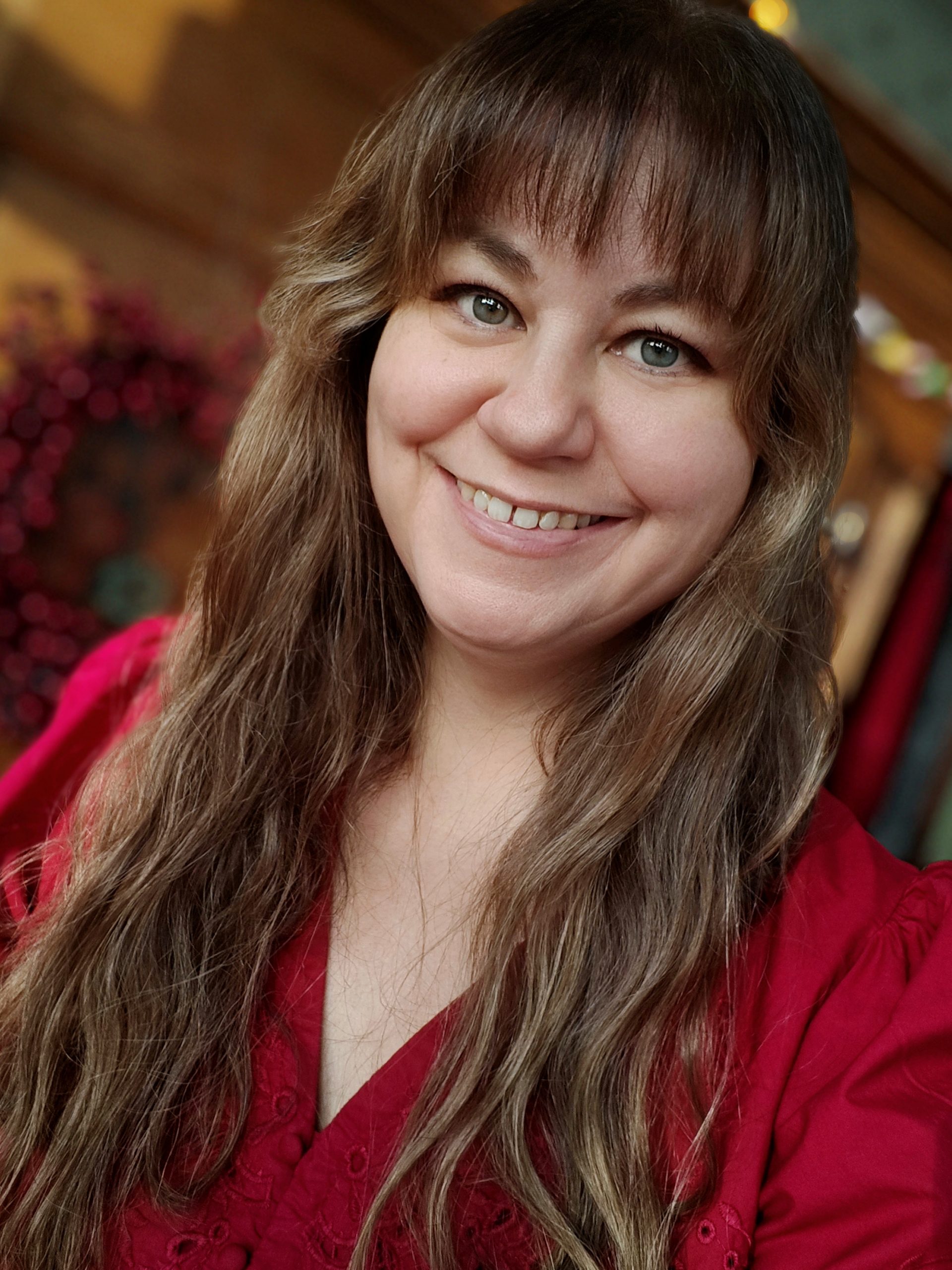 En kvinna med långt vågigt hår och lugg ler, klädd i en röd blus i ett varmt upplyst rum.