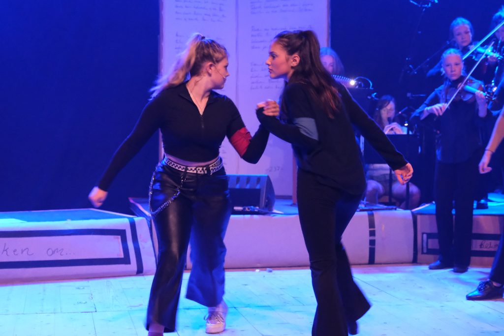 Två kvinnor håller hårt i varandras händer på scenen och verkar gräla under en dramatisk föreställning med musiker bakom sig.