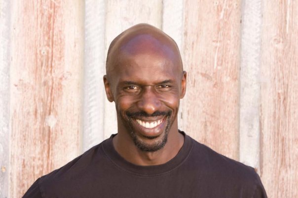 En leende man i svart skjorta står framför en ljus trävägg.