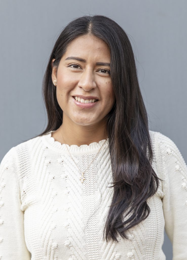 Kvinna med långt mörkt hår, klädd i vit tröja, ler mot en grå bakgrund.