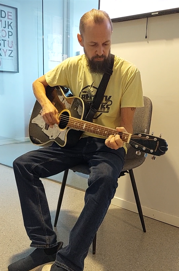 En man med skägg sitter på en stol och spelar akustisk gitarr i en modern inomhusmiljö.