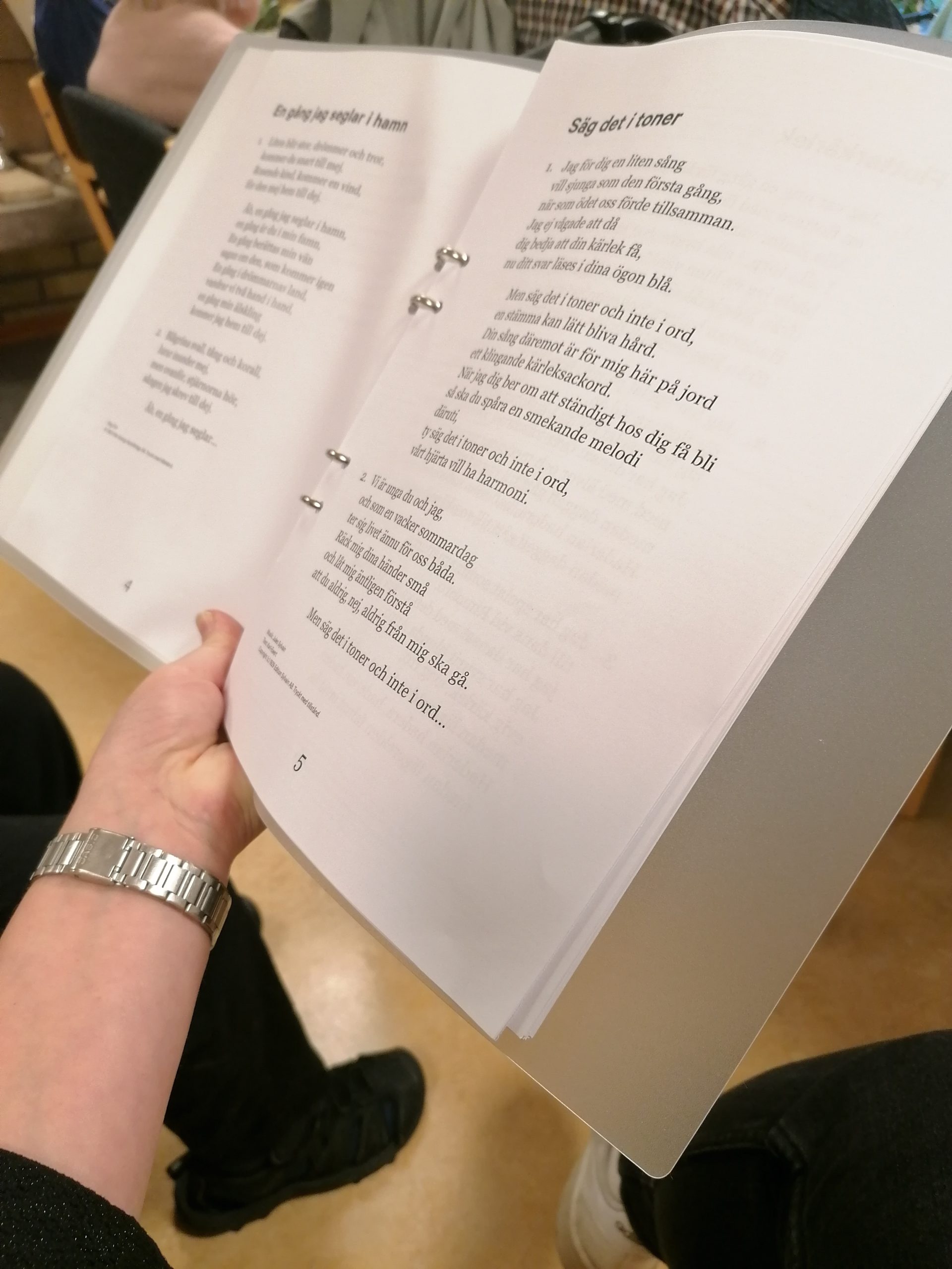 En person som håller i en öppen pärm med tryckt text, kanske poesi eller sångtexter, i ett klassrum.