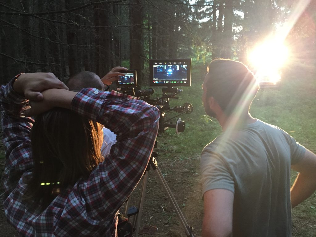 Två personer filmar en scen i en skog med hjälp av kameror och monitorer medan solljuset skiner genom träden.