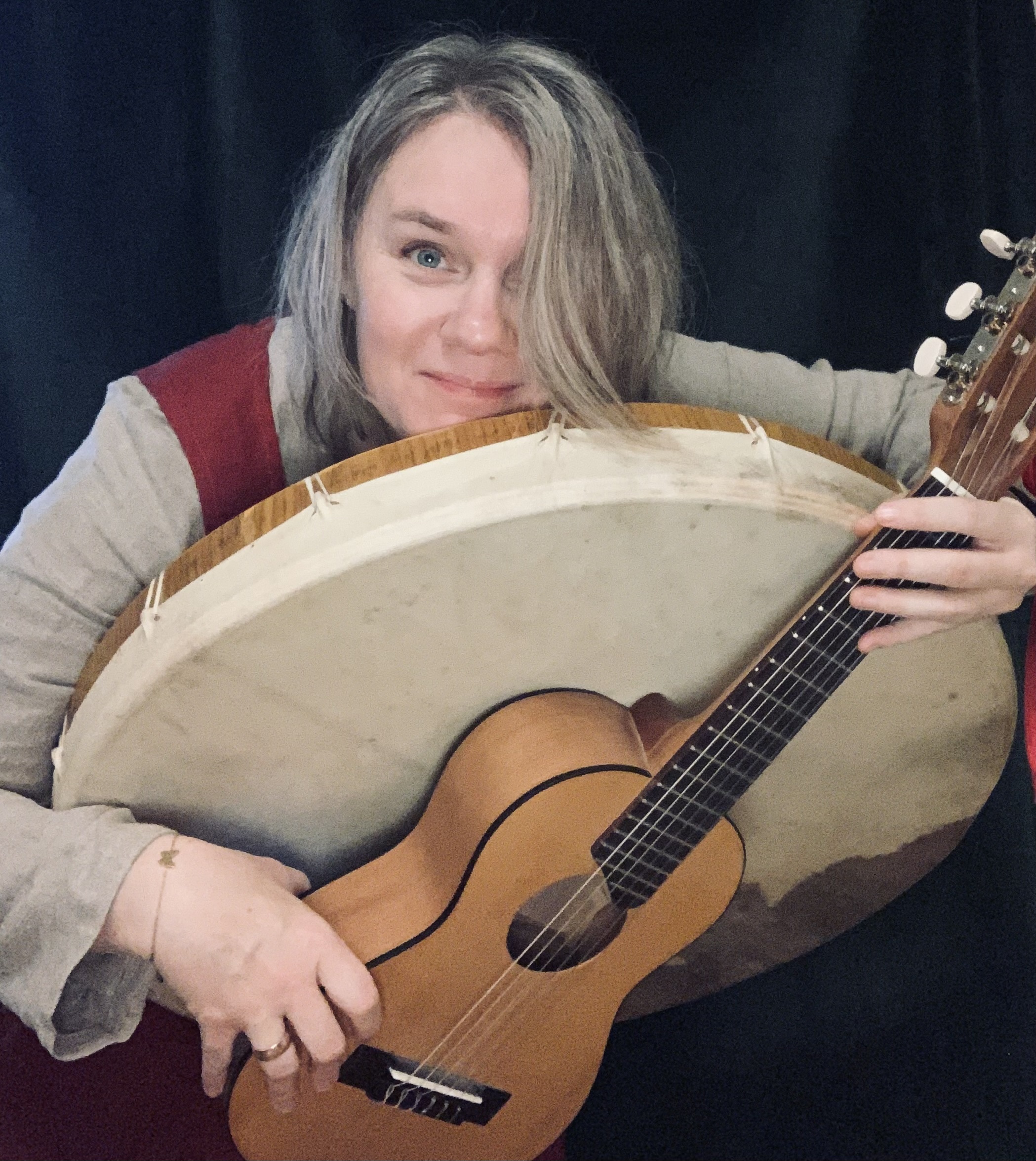 Leende person som håller i en stor ramtrumma och en akustisk gitarr mot en mörk bakgrund.