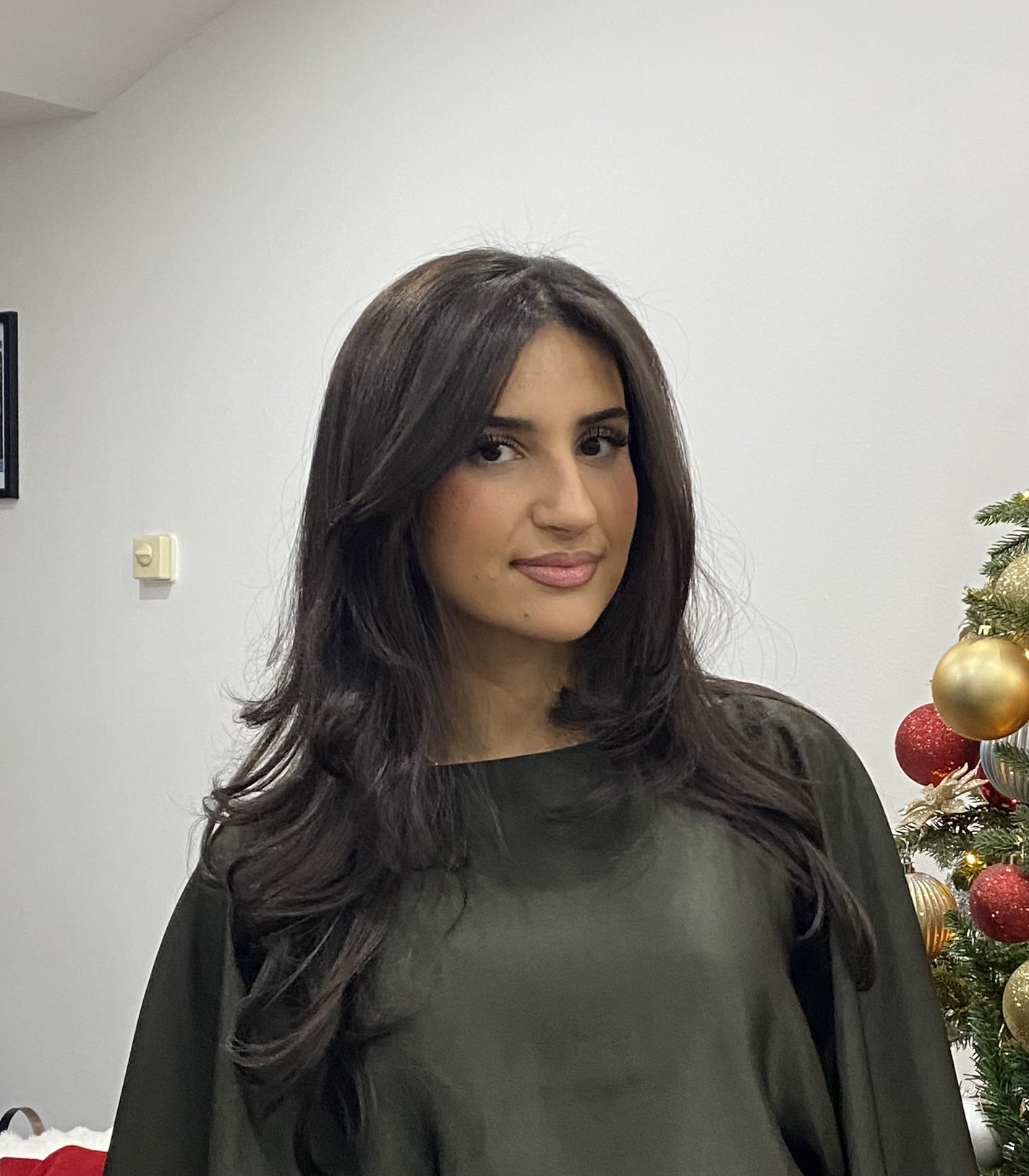 En kvinna med långt mörkt hår står inomhus nära en dekorerad julgran med guld- och röda ornament.