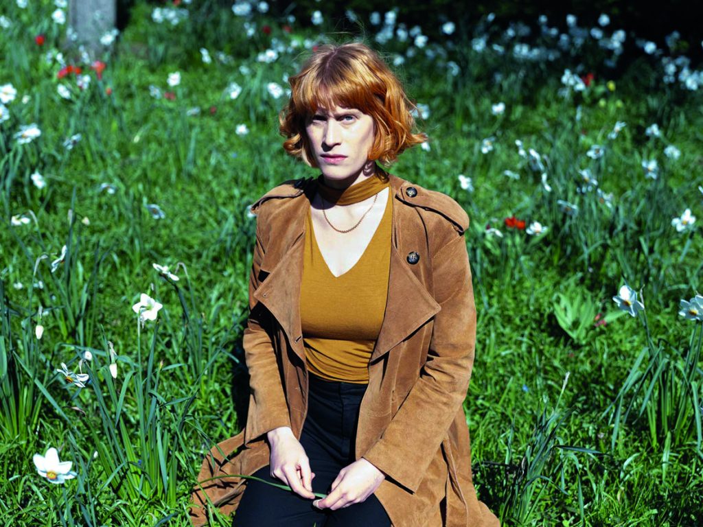 En kvinna med rött hår sitter på ett grässtrå omgivet av vita blommor, klädd i brun rock och senapsfärgad topp.