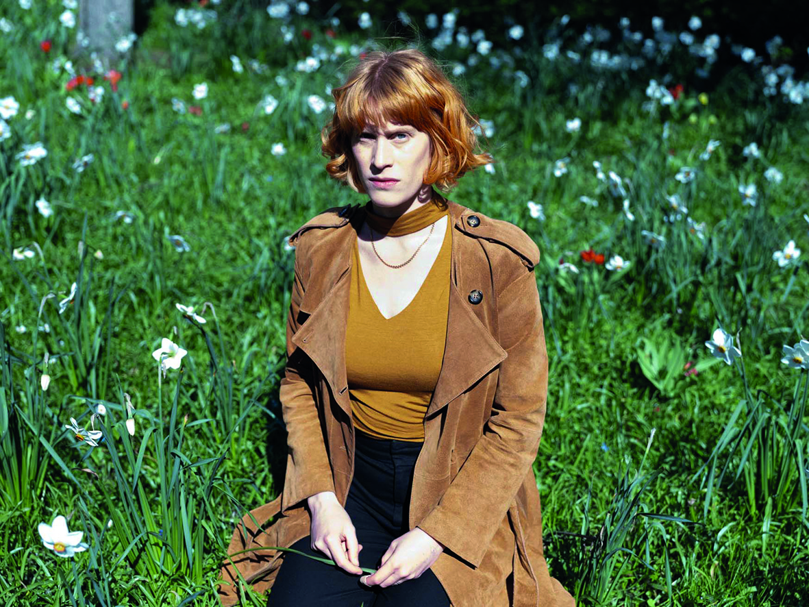 En kvinna med rött hår sitter på ett grässtrå omgivet av vita blommor, klädd i brun rock och senapsfärgad topp.