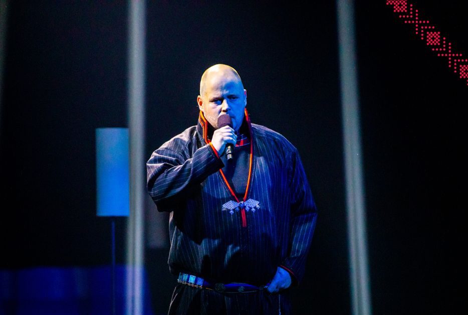 En man i traditionell klädsel talar i en mikrofon på scenen. Texten lyder "Jörgen Stenberg.