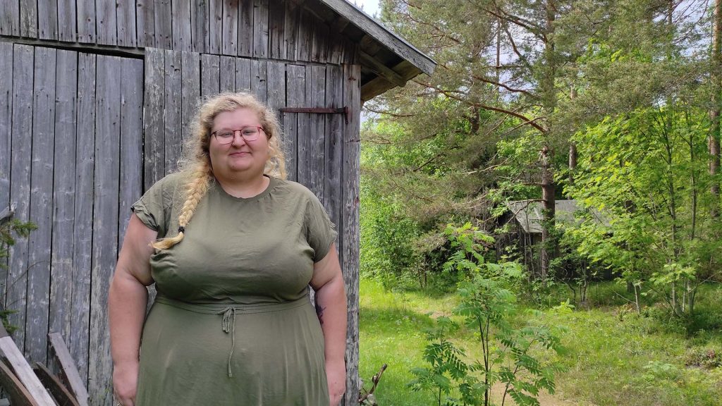 En kvinna med glasögon står vid ett väderbitet träskjul, med träd och grönska i bakgrunden.