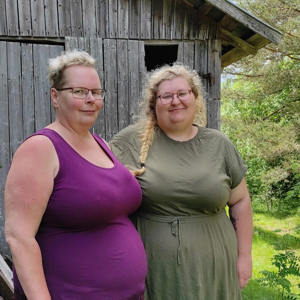 Två kvinnor står utanför ett träskjul, omgivna av grönska, och ler mot kameran.