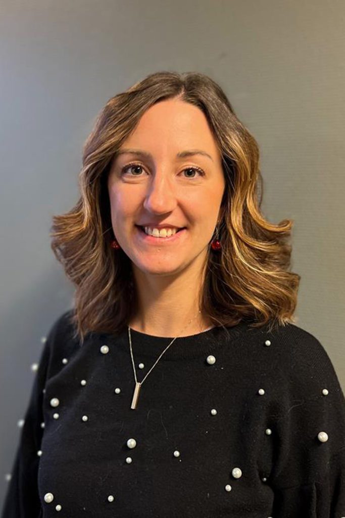 Kvinna med axellångt vågigt hår, bär en pärlbeströdd svart tröja och ett halsband, ler.