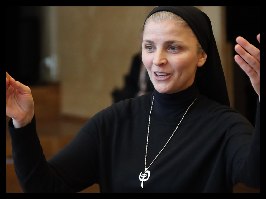 En kvinna med svart huvudduk och topp gestikulerar med båda händerna, ler och bär ett halsband.